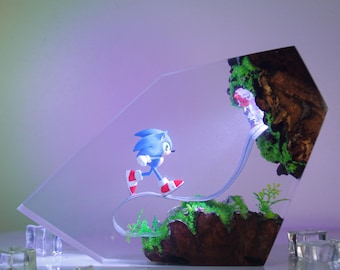 Könnte beinhalten: Eine blaue und weiße Sonic the Hedgehog Figur, die auf einem Weg in einer klaren Harzskulptur läuft. Die Skulptur zeigt eine mit grünem Moos bedeckte Felsformation und einen klaren Harz-Wasserfall.