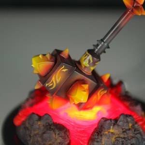 May include: A fiery, spiked hammer figurine with a glowing red base. The hammer is made of dark brown and orange plastic with yellow accents. The base is made of black plastic and has a red glow.