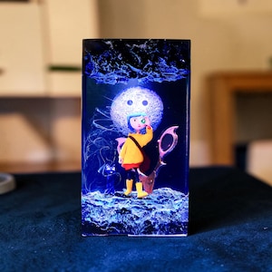 Puede incluir: Una pieza de arte de resina rectangular con un personaje de dibujos animados con abrigo amarillo, pantalones negros y botas amarillas. El personaje est&aacute; de pie frente a una luna brillante y un par de tijeras. La obra de arte tiene un fondo azul oscuro con acentos azules.