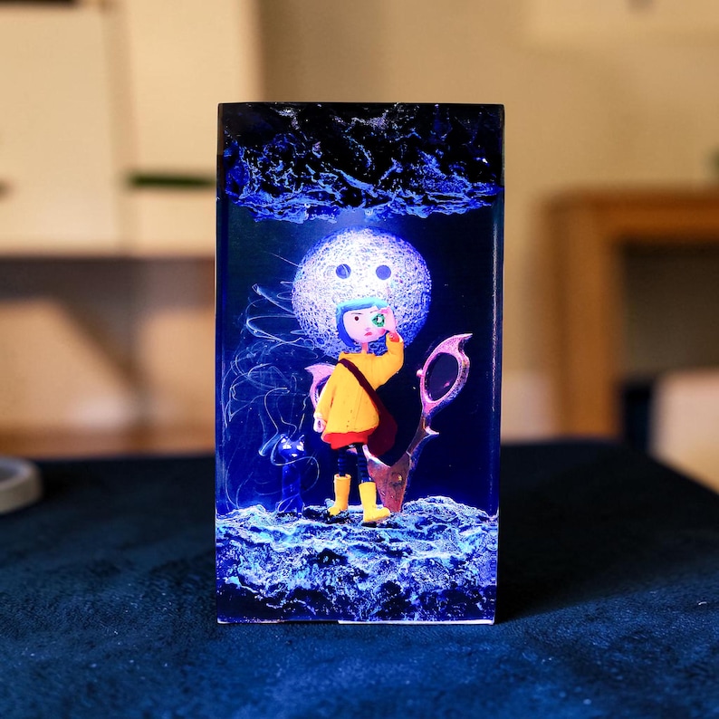 Puede incluir: Una pieza de arte de resina rectangular con un personaje de dibujos animados con abrigo amarillo, pantalones negros y botas amarillas. El personaje est&aacute; de pie frente a una luna brillante y un par de tijeras. La obra de arte tiene un fondo azul oscuro con acentos azules.