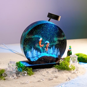 May include: A resin sculpture of two foxes leaping in front of a starry night sky. The sculpture is illuminated from below, creating a glowing effect. The sculpture is on a black base and surrounded by green moss and ice.