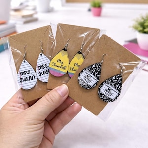 May include: Teardrop-shaped earrings with various designs, including lined paper with "Mrs. Susan", a pencil, and a composition notebook. Each pair is packaged on a brown card. The earrings are held in a person's hand.