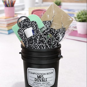May include: A black bucket filled with school supplies, including a composition notebook-themed keychain, earrings, and a lip balm. The bucket has a label that reads "COMPOSITION BOOK MRS. DOVALE" and measurements of 9 x 7 inches.