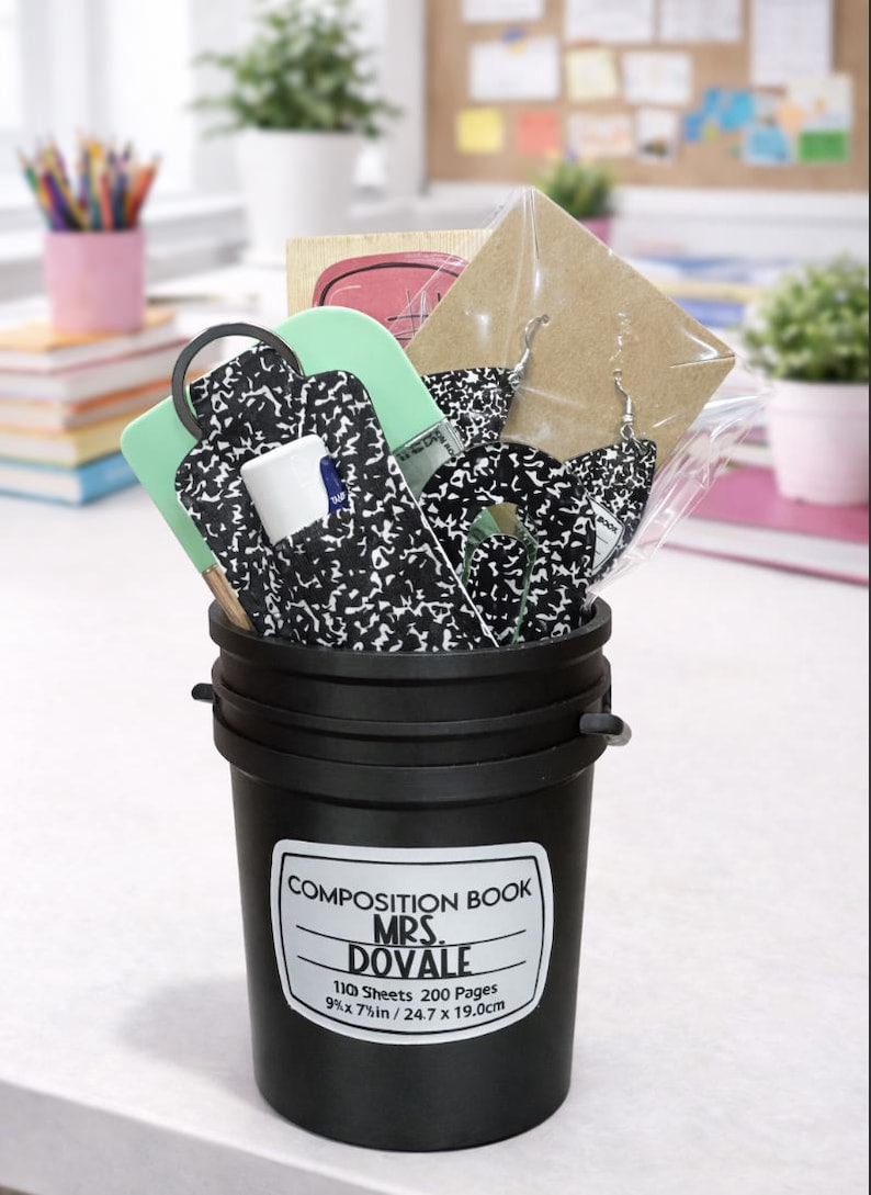 May include: A black bucket filled with school supplies, including a composition notebook-themed keychain, earrings, and a lip balm. The bucket has a label that reads "COMPOSITION BOOK MRS. DOVALE" and measurements of 9 x 7 inches.