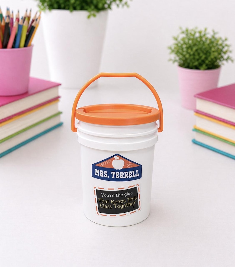 May include: White bucket with an orange lid and handle, featuring a label that reads "MRS. TERRELL" and "You're the glue That Keeps This Class Together." The bucket is surrounded by school supplies, including books and pencils.