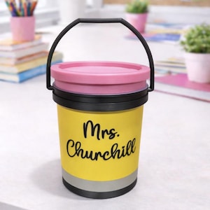 May include: A yellow pencil-shaped container with a black handle and lid. The container has the name "Mrs. Churchill" in black script. The lid is pink, and the container has a silver band near the bottom. The container is on a white surface.