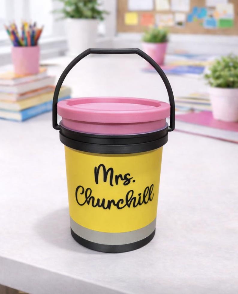 May include: A yellow pencil-shaped container with a black handle and lid. The container has the name "Mrs. Churchill" in black script. The lid is pink, and the container has a silver band near the bottom. The container is on a white surface.
