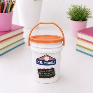 May include: White bucket with an orange lid and handle, featuring a label that reads "MRS. TERRELL" and "You're the glue That Keeps This Class Together." The bucket is surrounded by school supplies, including books and pencils.