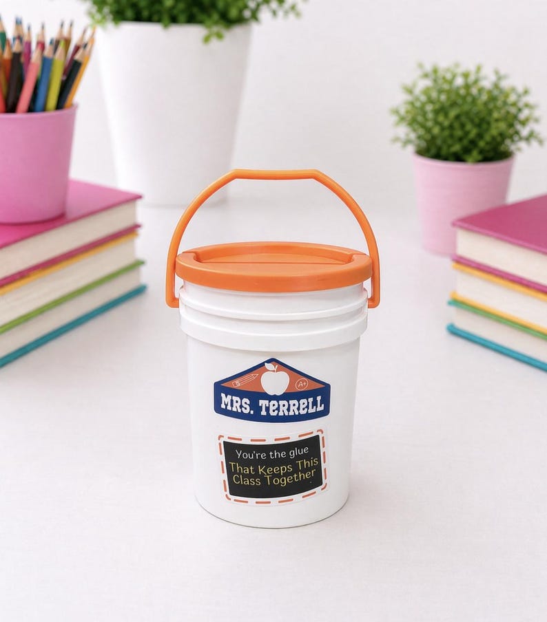 May include: White bucket with an orange lid and handle, featuring a label that reads "MRS. TERRELL" and "You're the glue That Keeps This Class Together." The bucket is surrounded by school supplies, including books and pencils.