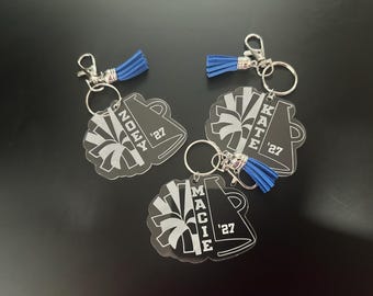 May include: Three clear acrylic keychains featuring a megaphone and pom-pom design with blue tassels. Each keychain is personalized with a name, NOEY, KATE, or MACIE, and the year '27 engraved in white. They include silver metal rings and clasps.