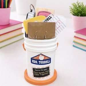 May include: A white bucket with orange handles and base, filled with gifts. The bucket has a label that reads "MRS. TERRELL" and "You're the glue That Keeps This Class Together." Inside are earrings, keychains, and other small items.