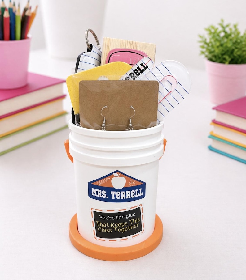 May include: A white bucket with orange handles and base, filled with gifts. The bucket has a label that reads "MRS. TERRELL" and "You're the glue That Keeps This Class Together." Inside are earrings, keychains, and other small items.