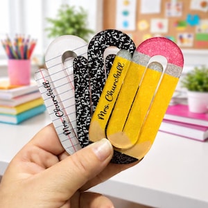 May include: A hand holds a collection of decorative bookmarks. The bookmarks feature designs like lined paper, a black and white composition notebook pattern, and a yellow pencil with a pink eraser. Each bookmark is labeled "Mrs. Churchill".