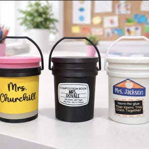 May include: Three small, bucket-shaped containers with black handles. One is yellow with "Mrs. Churchill" on it, another is black with "Composition Book Mrs. Dovale" on it, and the last is white with "Mrs. Jackson" on it.