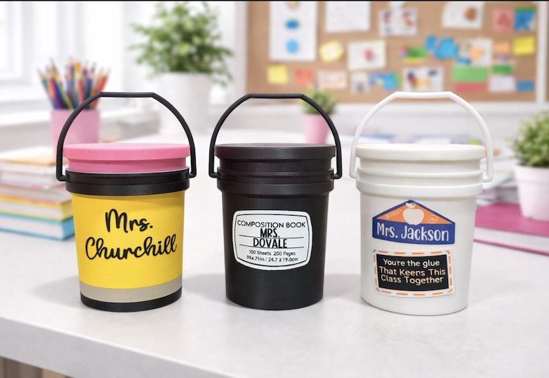 May include: Three small, bucket-shaped containers with black handles. One is yellow with "Mrs. Churchill" on it, another is black with "Composition Book Mrs. Dovale" on it, and the last is white with "Mrs. Jackson" on it.