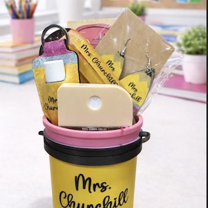 May include: A yellow pencil-themed gift bucket with the name "Mrs. Churchill" on the side, filled with various teacher-themed gifts. Includes a keychain, earrings, and a wooden ruler. The bucket has a pink rim and black handle.