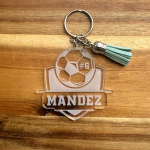 May include: Clear acrylic soccer keychain with a soccer ball design, the number 6, and the name "MANDEZ". The keychain has a silver keyring, chain, and a light teal tassel.