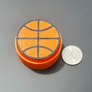 May include: An orange, 3D-printed container with a basketball design on top. The basketball graphic is orange with black lines. A U.S. quarter coin is next to the container for size comparison.