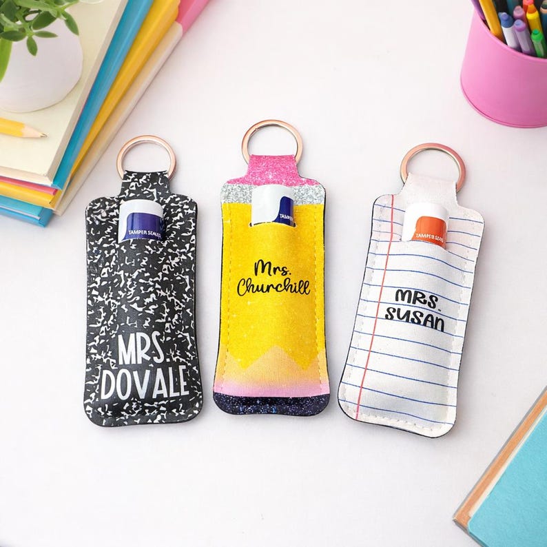 May include: Three personalized lip balm holders with key rings. One is black and white with "Mrs. Dovale" printed on it. The second is yellow with "Mrs. Churchill" and a pencil design. The third is white with blue lines and "Mrs. Susan".