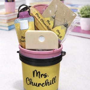 May include: A yellow pencil-themed gift bucket with a pink rim and black handle, filled with school-themed gifts. The bucket has the name "Mrs. Churchill" in black script. Inside are pencil-shaped earrings, a keychain, and a small, off-white item.