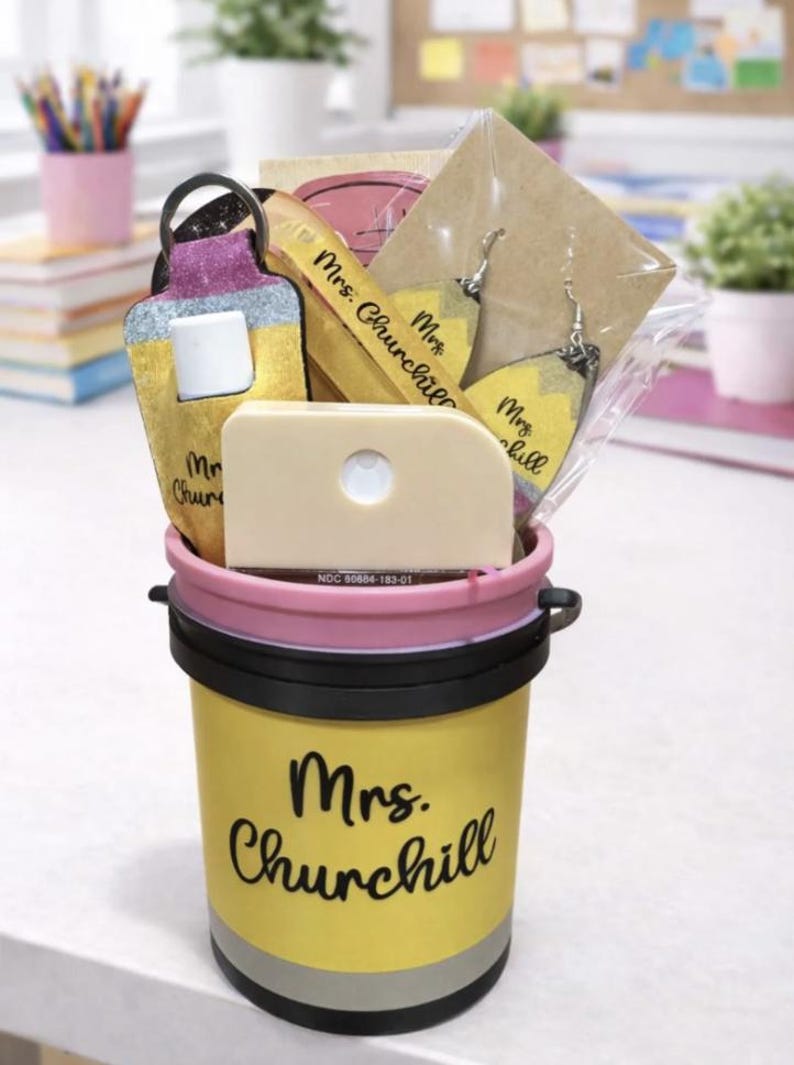 May include: A yellow pencil-themed gift bucket with a pink rim and black handle, filled with school-themed gifts. The bucket has the name "Mrs. Churchill" in black script. Inside are pencil-shaped earrings, a keychain, and a small, off-white item.