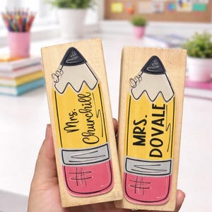 May include: Two wooden stamps with pencil designs. The pencils are yellow with pink erasers and gray tips. One stamp reads "Mrs. Churchill" and the other reads "Mrs. Dovale".