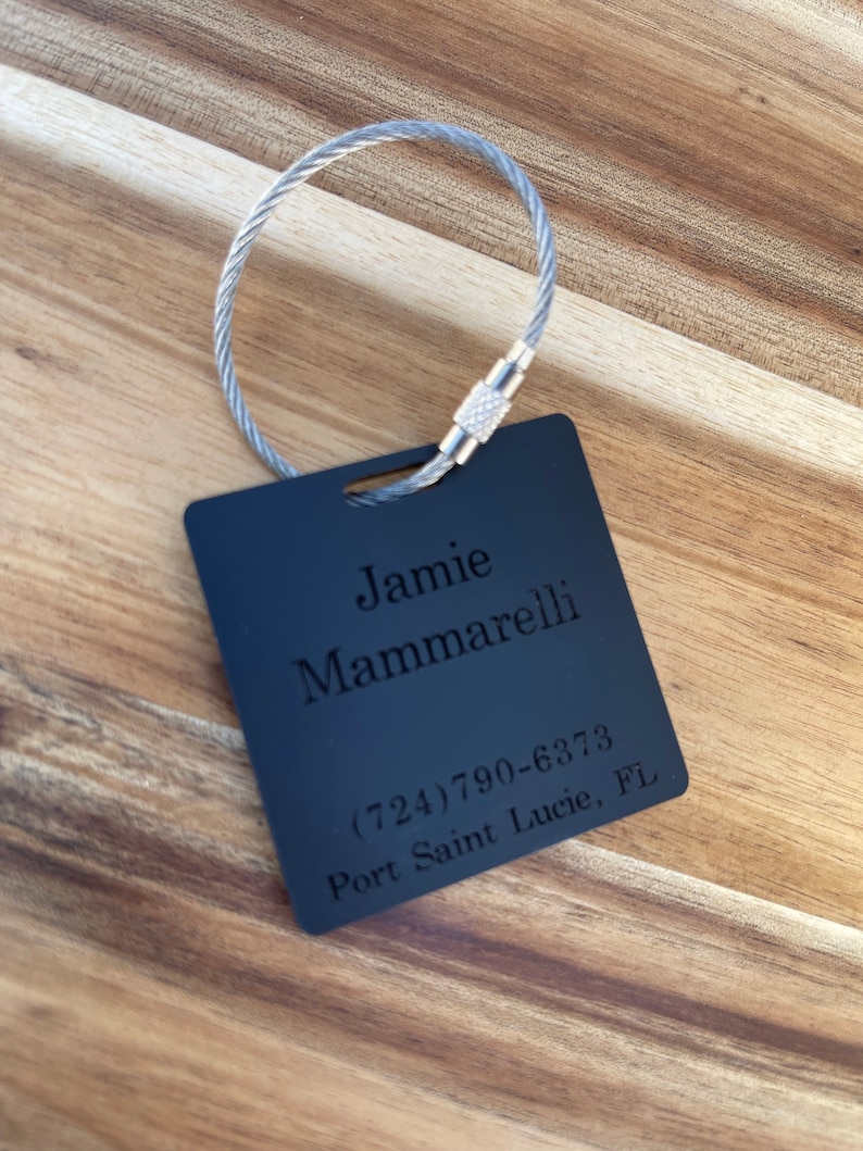 Puede incluir: Etiqueta de equipaje negra con un lazo de cable plateado. La etiqueta tiene el nombre Jamie Mammarelli, el n&uacute;mero de tel&eacute;fono (724) 790-6373 y la ubicaci&oacute;n Port Saint Lucie, FL.