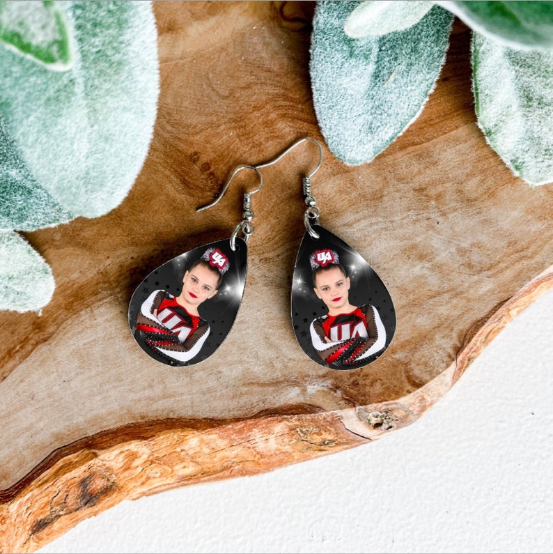 May include: A pair of teardrop-shaped earrings with a black background and a photo of a young girl wearing a red and white cheerleading uniform. The girl is wearing a headband with the number "14" on it. The earrings are silver.