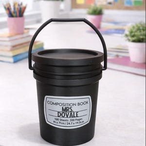 May include: Black bucket-shaped container with a handle, featuring a label that reads "COMPOSITION BOOK MRS. DOVALE." The label also indicates 100 sheets, 200 pages, and dimensions of 9 x 7 inches. The container is on a white surface.