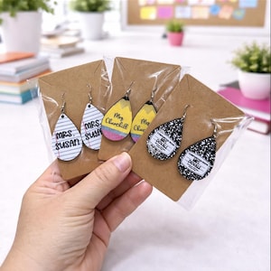 May include: Teardrop-shaped earrings with various designs, including lined paper with "Mrs. Susan", a pencil, and a composition notebook. Each pair is packaged on a brown card. The earrings are held in a person's hand.