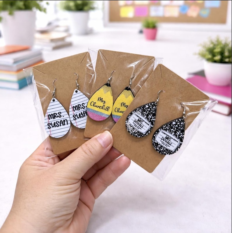 May include: Teardrop-shaped earrings with various designs, including lined paper with "Mrs. Susan", a pencil, and a composition notebook. Each pair is packaged on a brown card. The earrings are held in a person's hand.
