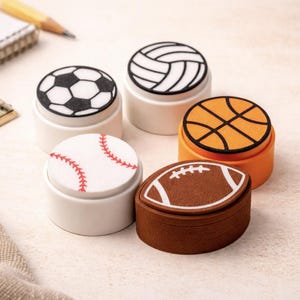 May include: A collection of sports-themed stamps featuring a soccer ball, volleyball, baseball, basketball, and football. The stamps have white bases with the sports designs in black, orange, and brown.