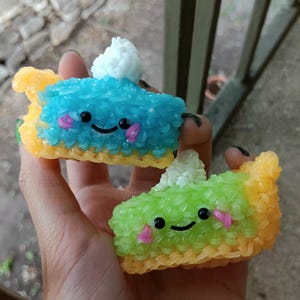 May include: Two colourful rubber band pie slice charms. One is blue, the other green, both with smiling faces, pink cheeks, and white whipped cream. The crusts are yellow and orange. These handmade charms are a fun accessory.