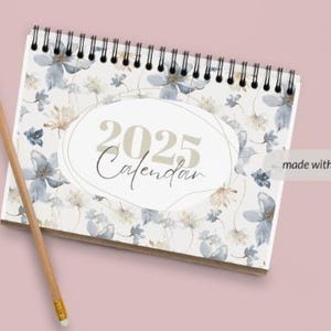 Könnte beinhalten: Ein weißer Spiralblockkalender mit einem floralen Design mit blauen und braunen Aquarellblumen. Der Kalender ist für das Jahr 2025 und trägt die Aufschrift "2025 Calendar" in einem goldenen Oval.