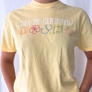 May include: A light yellow t-shirt with the embroidered text "SEA-PR CERTIFIED" above colorful seashell, flower, stethoscope, shell, and starfish designs. The shirt is paired with green and white checkered shorts.