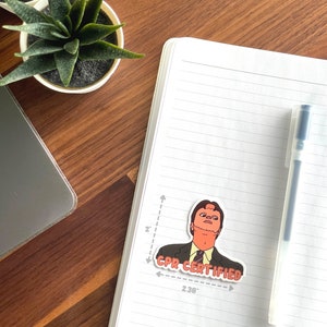 May include: A sticker featuring a cartoon character in a suit with the text "CPR CERTIFIED" in red. The sticker is approximately 2 inches tall and 2.38 inches wide. The sticker is on a white notebook with a pen.