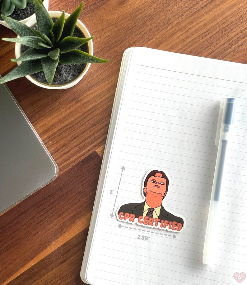May include: A sticker featuring a cartoon character in a suit with the text "CPR CERTIFIED" in red. The sticker is approximately 2 inches tall and 2.38 inches wide. The sticker is on a white notebook with a pen.