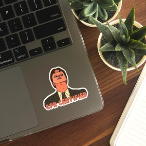 May include: A sticker featuring a cartoon image of a man in a suit with the text "CPR CERTIFIED" in a red and white banner. The sticker is affixed to a laptop, with two small potted succulents and a notebook in the background.