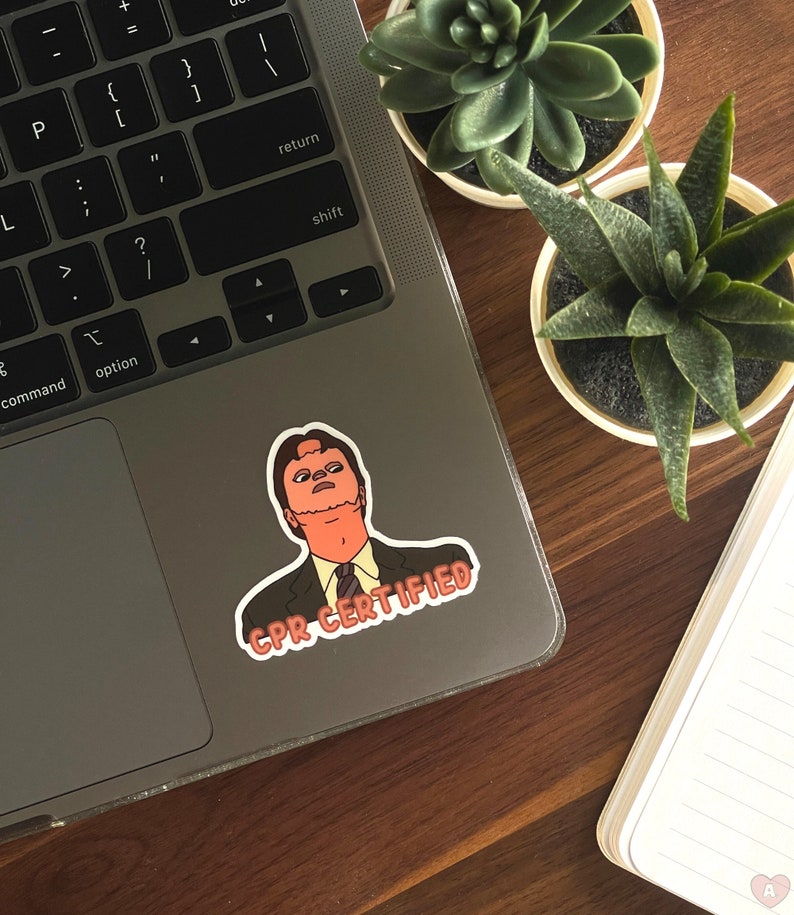 May include: A sticker featuring a cartoon image of a man in a suit with the text "CPR CERTIFIED" in a red and white banner. The sticker is affixed to a laptop, with two small potted succulents and a notebook in the background.
