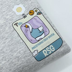 May include: A gray t-shirt with a cartoon goose wearing a blue shirt and holding a syringe. The goose is in a light blue frame with the text "Registered Silly Goose" and "RSG".