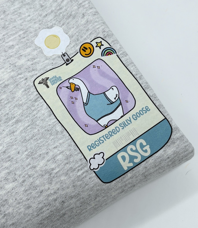 May include: A gray t-shirt with a cartoon goose wearing a blue shirt and holding a syringe. The goose is in a light blue frame with the text "Registered Silly Goose" and "RSG".
