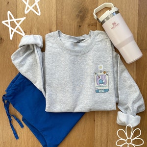 May include: A gray sweatshirt with a cartoon goose graphic and the text "patchworked soul patch" and "RSG". The sweatshirt is laid out on a wooden surface with a blue pair of pants and a pink Stanley water bottle.