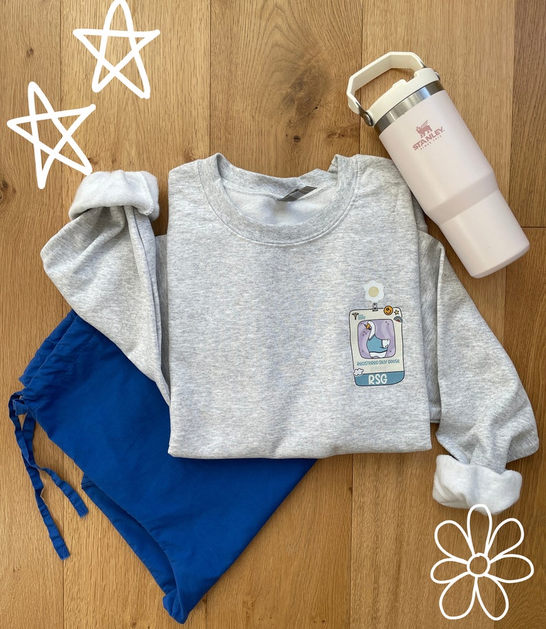 May include: A gray sweatshirt with a cartoon goose graphic and the text "patchworked soul patch" and "RSG". The sweatshirt is laid out on a wooden surface with a blue pair of pants and a pink Stanley water bottle.