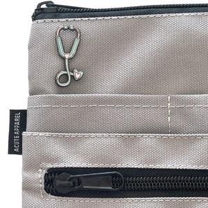 May include: A gray zippered pouch with a black zipper and a stethoscope pin with pink and blue accents. The pouch has the text "ACUTE APPAREL" printed on the side.