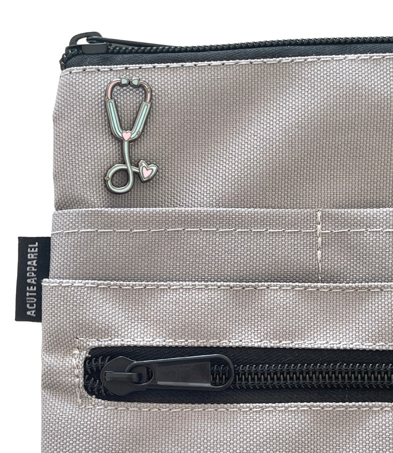 May include: A gray zippered pouch with a black zipper and a stethoscope pin with pink and blue accents. The pouch has the text "ACUTE APPAREL" printed on the side.