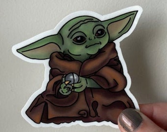 May include: A green and brown cartoon sticker of a character from the Star Wars franchise, known as "The Child" or "Baby Yoda", holding a small silver ball in its hand.