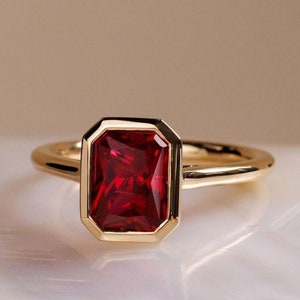 Pode incluir: Um anel de ouro com uma gema vermelha engastada em um bezel quadrado.