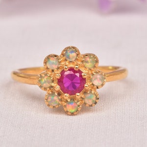 May include: A gold ring featuring a flower-shaped design. The center of the flower is a round, bright pink gemstone, surrounded by smaller, iridescent opal stones. The ring is set against a soft, white background.