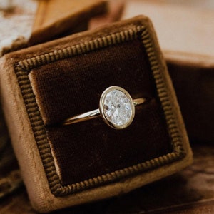May include: A gold ring with an oval-shaped diamond set in a bezel setting. The ring is displayed in a brown velvet ring box.