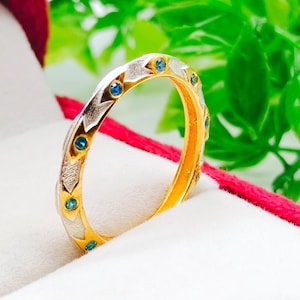May include: A gold ring with a silver band featuring blue gemstones. The ring is set in a red jewelry box with a white interior, against a backdrop of green foliage. The ring's design includes alternating silver and gold sections.